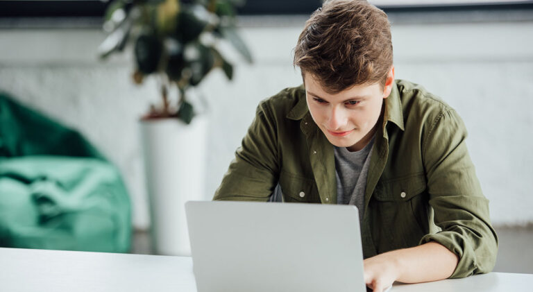 smiling teen boy sitting at table and using laptop